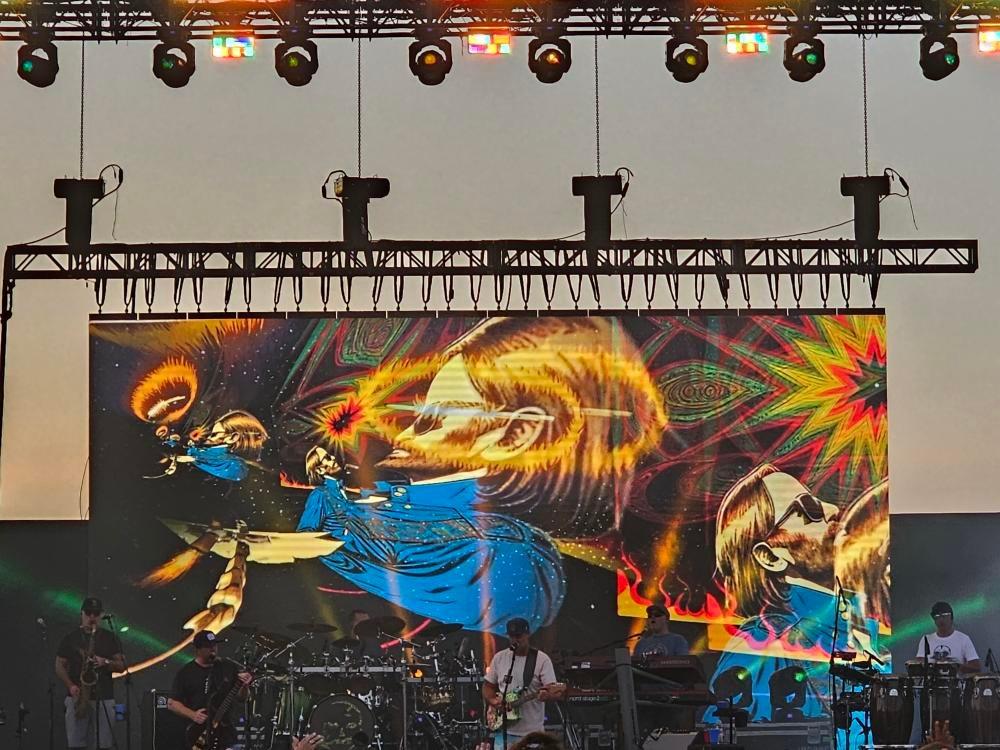 A luminous skull visual fills the screen above musicians on an outdoor stage.